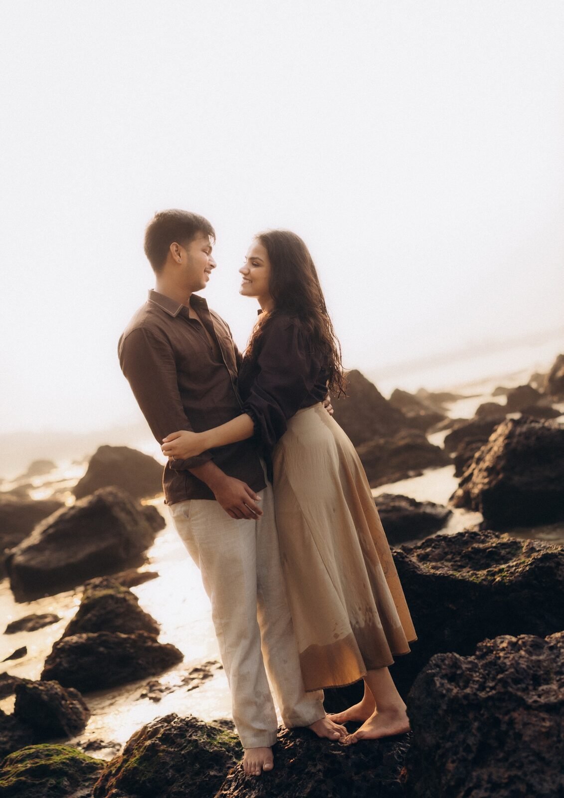 Couple standing on moss-covered rocks at the waterline in soft hazy light, Goa pre-wedding shoot