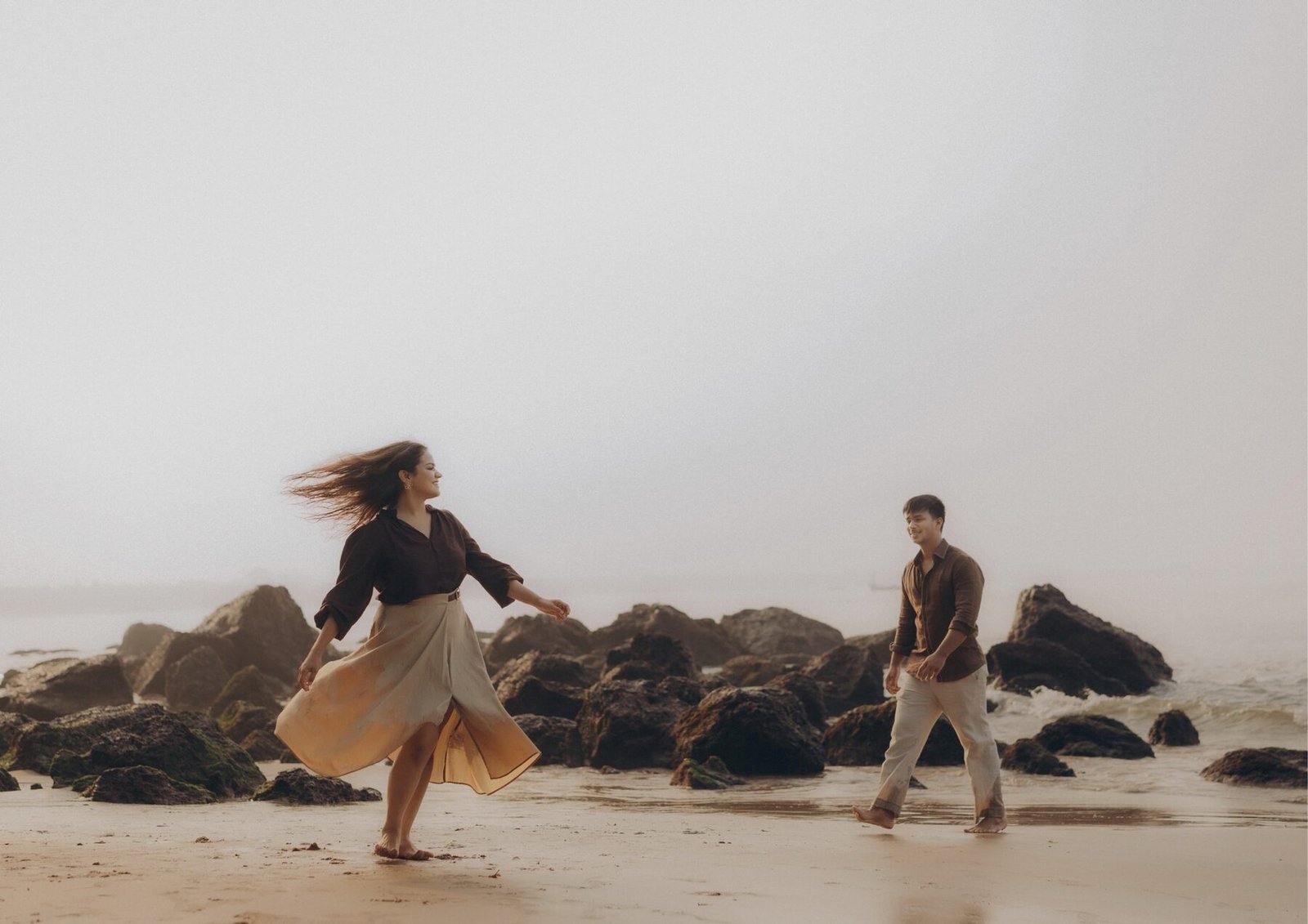 Wide frame of the couple on a misty rocky Goa beach, skirt caught by the wind